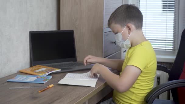 Un niño con una máscara médica está dando clases en la mesa durante la pandemia de Covid. — Vídeos de Stock