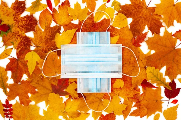 Protective medical face masks lie on maple, oak, birch leaves and form a medical cross. Autumn background. Protection against coronavirus infection covid-19. Flatly, top view