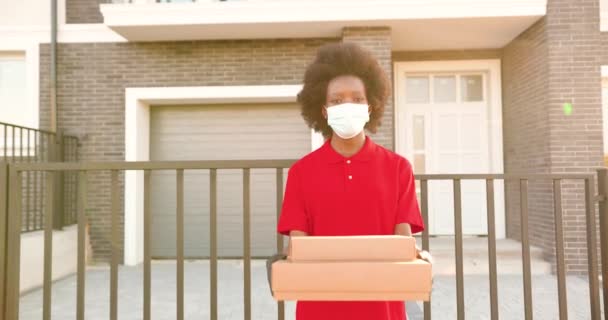 Egy portré afro-amerikai fiatal női futárról orvosi maszkban és kesztyűben, aki az utcán áll, és kartondobozokat tart a kezében. A női kézbesítő dobozokat ad. Karantén. Pizza átadása. — Stock videók