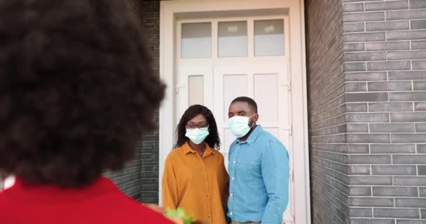 Achterzijde van Afro-Amerikaanse vrouwelijke koerier leveren tas met gezond voedsel aan paar thuis, overhandigen aan mannelijke outdoor. Bezorgster brengt pakje groente naar man en vrouw. Coronavirusconcept — Stockvideo