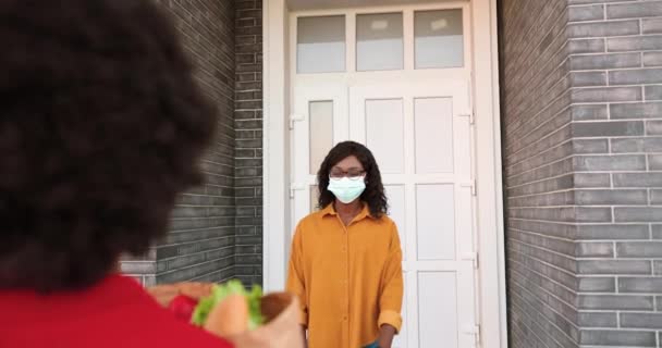 Afroamerykańska kurierka dostarczająca torbę ze zdrowym jedzeniem do klienta w domu i rozdająca na zewnątrz. Dostawczyni przynosząca paczkę warzyw do klienta. Koncepcja koronawirusa. Kwarantanna — Wideo stockowe