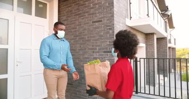 African American female courier in mask delivering bag with healthy food to male client at house and handing outside. Delivery woman bringing packet of vegetables to man customer. Coronavirus concept. — Stock Video