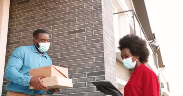 Coursier afro-américain femme en masque livrant des colis en carton à un client masculin à la maison et la remise à l'extérieur. Livraison femme apportant des boîtes de pizza à l'homme client. Concept de coronavirus. — Video