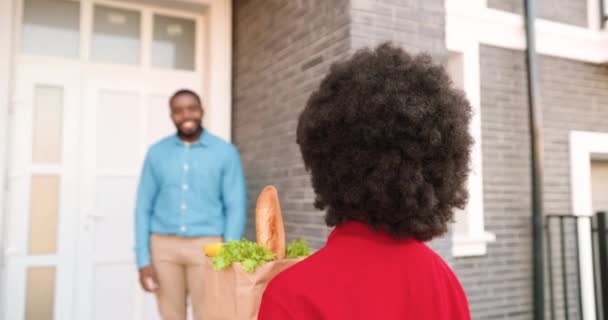 Αφρο-Αμερικανίδα κούριερ παραδίδει σακούλες με υγιεινό φαγητό σε άντρα πελάτη στο σπίτι, μιλώντας και μοιράζοντας έξω. Παράδοση γυναίκα χαμογελώντας και φέρνοντας πακέτο των λαχανικών στον άνθρωπο πελάτη. Πίσω. — Αρχείο Βίντεο