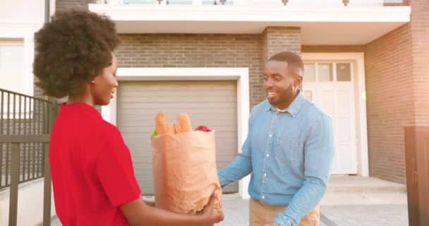 Αφρο-Αμερικανίδα κούριερ παραδίδει τσάντες με φαγητό σε άντρα πελάτη στο σπίτι, μιλώντας και μοιράζοντας έξω. Παράδοση γυναίκα χαμογελώντας και φέρνοντας πακέτο των προϊόντων στον άνθρωπο πελάτη. — Αρχείο Βίντεο