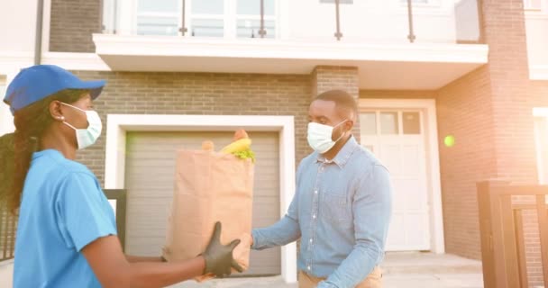 Correio feminino afro-americano em máscara médica entregando saco com comida para cliente masculino em casa e entregando fora. Mulher de entrega trazendo pacote de produtos para o homem cliente. Bloqueio do coronavírus. — Vídeo de Stock