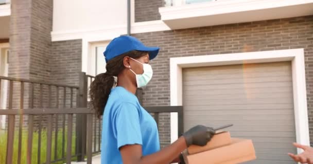 Tıbbi maskeli Afro-Amerikan kadın kurye evdeki erkek müşterilere karton paketler dağıtıyor ve dışarıda dağıtıyor. Kargocu kadın kutuları erkeğe getiriyor ve tablet cihazıyla karantina yapıyor. — Stok video