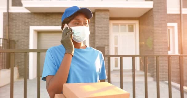 Афроамериканська молода жінка в медичній масці, рукавицях і капелюсі стоїть на вулиці з картонними посилками і розмовляє по мобільному телефону. Жінка - кур "єр у формі говорить по мобільному телефону. — стокове відео