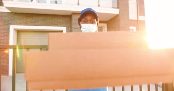 Молодая афроамериканка в медицинской маске, перчатках и шляпе стоит на улице с коробками посылок и вручает камеру. Курьер в форме раздает коробки. Предоставление концепции пандемии. — стоковое видео