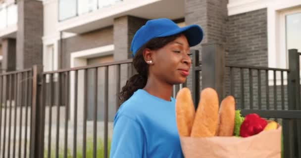 La mensajera afroamericana entrega la bolsa con comida al cliente en casa, sonriendo y entregando afuera. Entrega mujer sonriendo y trayendo paquete de productos para niña cliente. Día de verano. — Vídeo de stock