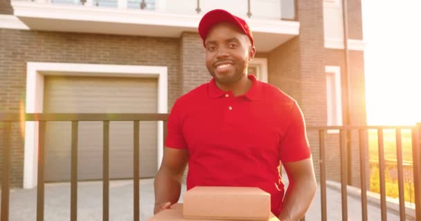 Portrait of smiled African American young man courier in red uniform and hat standing at house in street and holding carton parcels. Pekerja pengiriman laki-laki berseragam dengan kotak. tersenyum dengan gembira. — Stok Video