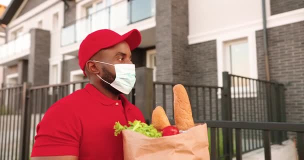 Mensajero masculino afroamericano en máscara médica entrega bolsa con comida a cliente femenino en casa y entrega al aire libre. Entrega hombre trayendo paquete de productos a la mujer cliente. Bloqueo de Corona. — Vídeo de stock