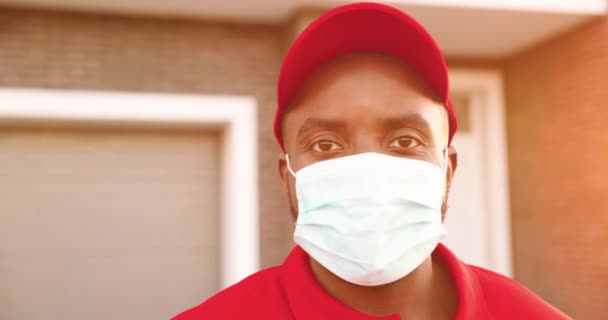 Retrato de jovem afro-americano feliz em máscara médica em pé fora da casa no dia de verão. Feche de mensageiro alegre masculino no uniforme. Entregador com sorriso na rua. Conceito de pandemia. — Vídeo de Stock