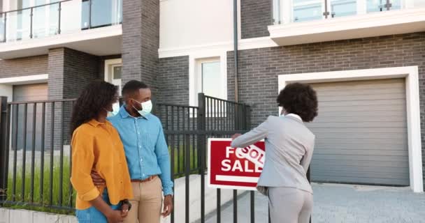 African American young woman real-eastate agent selling house to happy just-married couple. Outside. Changing board for sale to sold. Wealthy man and woman buying dwelling in suburb. Coronavirus time. — Stock Video