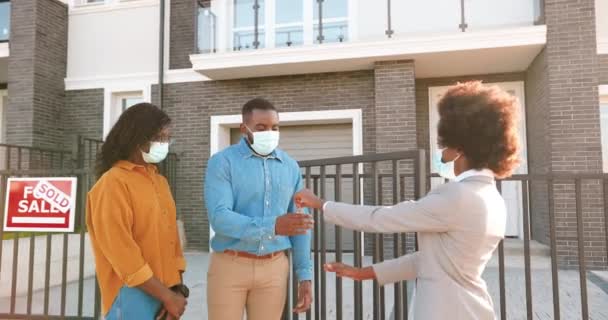 Female real-estate selling house to African American happy couple and passing keys. Outdoor. Cheerful just-married man and woman in medical masks hugging when buying home at outskirt. Covid-19 concept — Stock Video