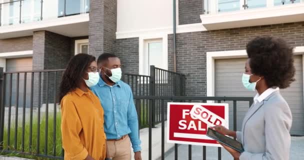 Afro-Américain jeune couple heureux dans des masques médicaux acheter une maison à la périphérie et de parler avec l'agent immobilier féminin tenant tablette dispositif dans les mains. En plein air. Concept de pandémie de coronavirus. — Video