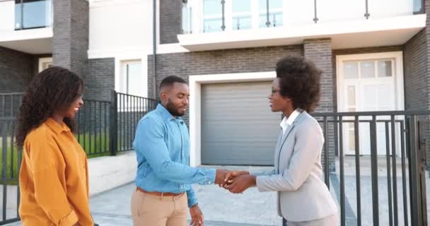 Inmobiliaria femenina vendiendo casa a pareja afro-americana feliz y pasando llaves. Al aire libre. Alegre hombre y mujer recién casados sonriendo y comprando casa en las afueras. Conseguir vivienda en el suburbio. — Vídeo de stock