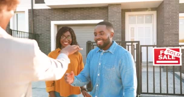 Afrikalı Amerikalı mutlu çifte ev satıp anahtarları teslim eden emlak şirketinin arkasında. Açık havada. Neşeli evli erkek ve kadın kucaklaşıp, kenar mahalleden ev alıyorlar. Banliyöde yaşamak. arka plan. — Stok video