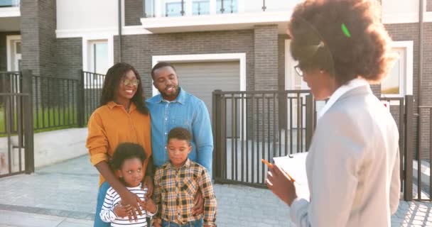 Happy African American family with two small kids buying house at suburbs and talking with female real-estate agent outdoors. Married couple with children looking for dwelling in outskirt neighborhood — Stock Video