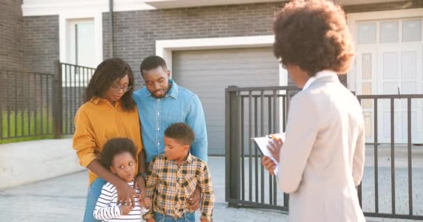 Jovem família afro-americana com duas crianças pequenas comprando casa nos subúrbios e conversando com agente imobiliário feminino ao ar livre. Casal casal com filhos à procura de habitação no bairro outskirt — Vídeo de Stock