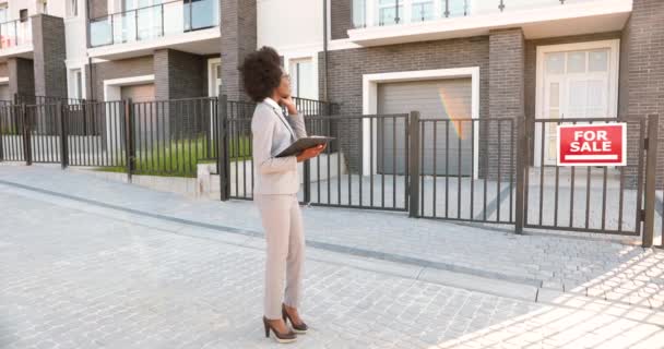 Afroamericana joven agente de bienes raíces de pie en la calle en el barrio suborbe y hablando por teléfono móvil. Empresaria con documentos buscando casa y hablando por celular. — Vídeo de stock