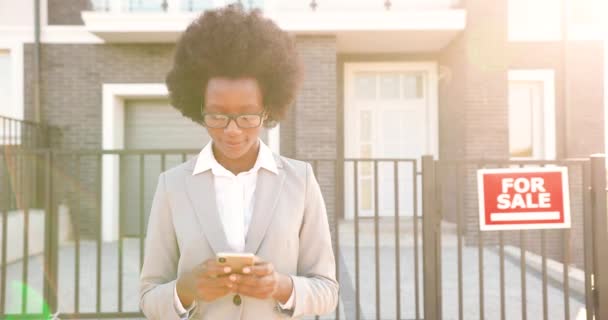 Jeune afro-américaine belle agent immobilier féminin élégant dans les lunettes et le style d'affaires debout en plein air dans la banlieue à la maison à vendre et SMS message sur smartphone. Utilisation du téléphone mobile. — Video