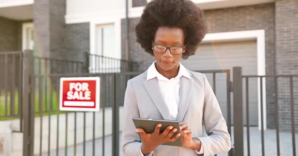 Νεαρή Αφροαμερικανίδα όμορφη γυναίκα κτηματομεσίτης σε γυαλιά και επιχειρηματικό στυλ στέκεται εξωτερική στο προάστιο στο σπίτι προς πώληση και γραπτών μηνυμάτων στη συσκευή tablet. Χρήση υπολογιστή gadget και πατώντας. — Αρχείο Βίντεο