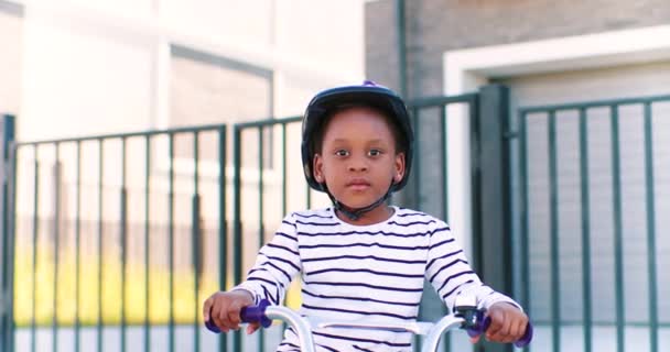 Портрет маленькой афроамериканской симпатичной девушки в каске, сидящей на велосипеде и улыбающейся в камеру на открытом воздухе. Маленький красивый ребенок в шлеме катается на велосипеде по улице в пригороде. — стоковое видео