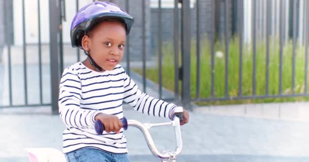 Портрет маленькой веселой афроамериканской милой девушки в каске, сидящей на велосипеде и улыбающейся в камеру на открытом воздухе. Маленький счастливый красивый ребенок в шлеме катается на велосипеде по улице на окраине. — стоковое видео