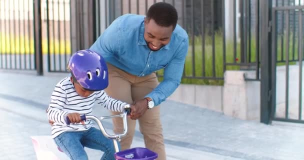 Красавчик-афроамериканец учит маленькую симпатичную девушку в шлеме кататься на велосипеде на улице в пригороде. Малыш ездит на велосипеде и учится. Школьница с папой. Дочь сидит на транспортном средстве — стоковое видео