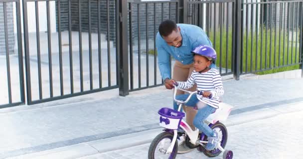 Afro-Amerikaanse knappe vader geeft les aan een klein mooi meisje in een helm op de fiets op straat in een buitenwijk. Een klein kind dat fietst en leert. Schoolmeisje bij papa. Dochter en vader. Ouderschap — Stockvideo