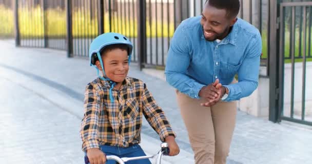 アフリカ系アメリカ人のハンサムな父親は、郊外の通りで自転車に乗ってヘルメットで小さな男の子を教えています。小さな子供が自転車に乗って学びます。パパと一緒にいる小学生。陽気な息子は車の上に座って. — ストック動画