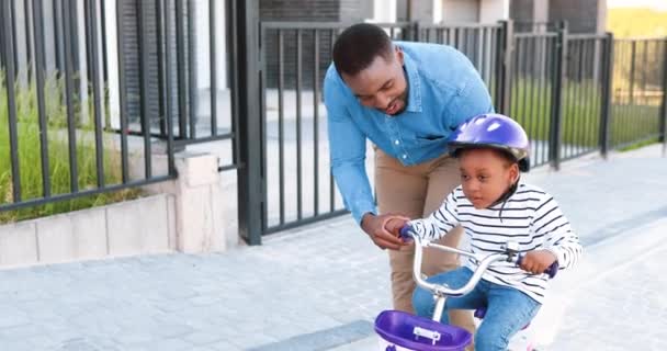 Afro-americano bonito pai feliz ensinando menina pequena no capacete andando de bicicleta na rua em saia. Um miúdo a andar de bicicleta e a aprender. Filha alegre com o pai. Fim de semana parental. — Vídeo de Stock
