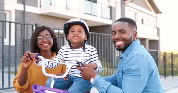 Πορτρέτο πλάνο της νεαρής Αφρικής αμερικανική ευτυχισμένη οικογένεια με μικρό χαριτωμένο κορίτσι στο ποδήλατο χαμογελώντας στην κάμερα εξωτερική. Χαρούμενη χαμογέλασε μητέρα και ο πατέρας με μικρή κόρη στο ποδήλατο στο δρόμο στο προάστιο. — Αρχείο Βίντεο