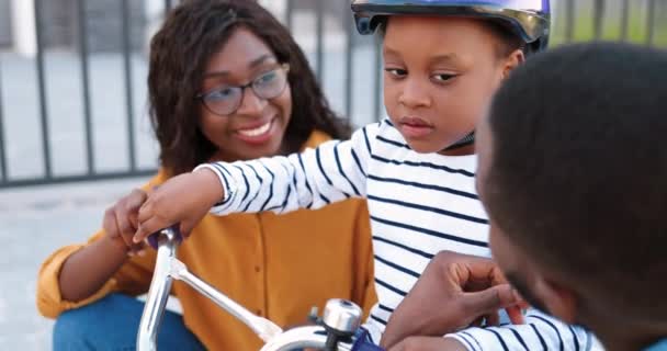 Primer plano de la pequeña chica linda afroamericana en casco sentado en bicicleta y escuchando a sus padres sonrientes. Alegre madre y padre enseñando bicicleta bonita hija. Montar en bicicleta. — Vídeos de Stock