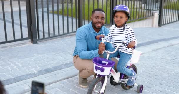 Οπίσθια Αφρο-αμερικανική μητέρα λήψη φωτογραφιών με κάμερα smartphone του συζύγου και της κόρης, ο άνθρωπος και μικρό χαριτωμένο κορίτσι με ποδήλατο. Πίσω όψη για γυναίκα λήψη φωτογραφιών στο κινητό τηλέφωνο του πατέρα και του παιδιού με ποδήλατο — Αρχείο Βίντεο