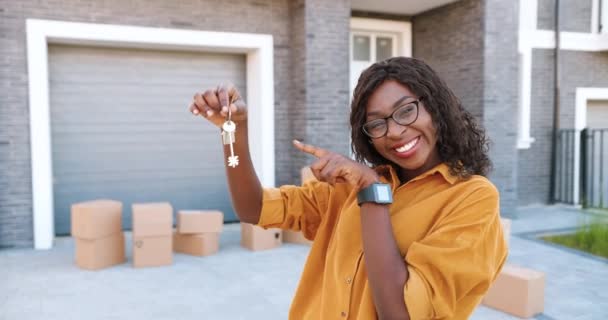 Retrato de alegre mujer afroamericana feliz en gafas sonriendo a la cámara y mostrando la clave de la cámara mientras se mueve en un nuevo hogar. Al aire libre. Hembra demostrando las llaves. Cajas de cartón sobre fondo. — Vídeo de stock