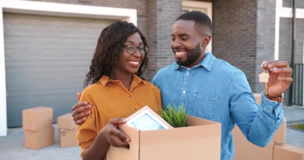 Retrato de una joven afroamericana casada pareja de hombres y mujeres de pie en un nuevo hogar, sosteniendo una caja de cartón con cosas y mostrando la llave al mudarse. Esposa y marido en el patio de la compra de bienes raíces. — Vídeos de Stock