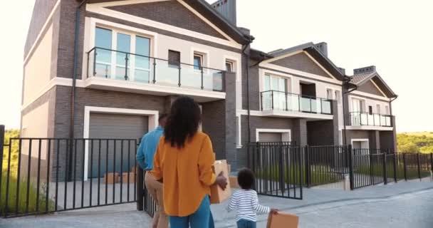 Zadní část afroamerické rodiny s malými dětmi na procházce a stěhování do nového domova na předměstí. Matka a otec nosí krabice v domě a malé děti pomáhají. Rodiče, syn a dcera se usazují. — Stock video