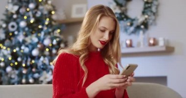 Güzel, neşeli, beyaz bir kadının portresi modern bir odada, Xmas ağacıyla akıllı telefondan mesaj atıyor ve Noel arifesinde selamlarını yolluyor. Şenlik havası. Xmas ruhu kavramı