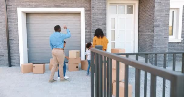 Rear on joyful African American parents with little children ordering carton boxes at yard of new home. Family with small kids moving in big house at outskirt. Settlement at subburb. Outdoors. — Stock Video