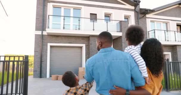 Trasera en la familia joven afroamericana con niños pequeños de pie en el patio de la casa y el padre señalando con el dedo en el segundo piso. Vista posterior de los padres con niños que se mudan a un nuevo hogar. — Vídeo de stock