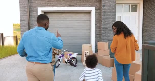 Šťastná afroamerická rodina s malými dětmi, které se stěhují do nového domova na předměstí. Matka a otec odnášejí krabice na dvorek a malé děti pomáhají. Rodiče se synem, dcerou a psem. — Stock video