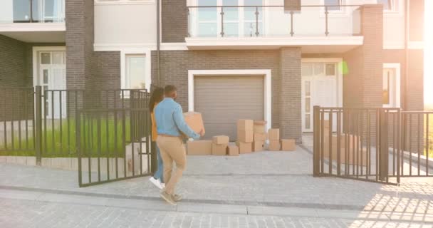 Šťastný pár Afroameričanů se stěhuje do nového domova na předměstí. Žena a muž nesoucí krabice na dvorek v domě a usazující se. Vyrovnání. Zadek. Zadní pohled. — Stock video