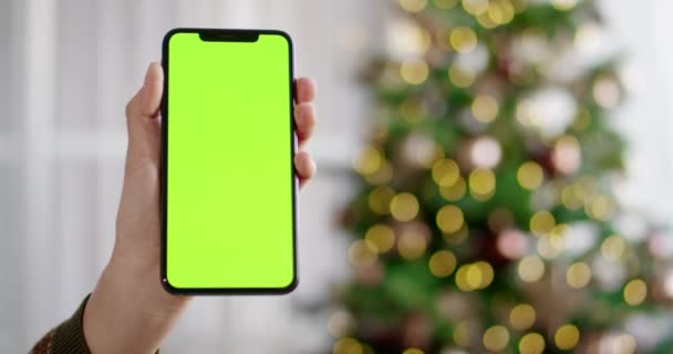 Primer plano de mano caucásica sosteniendo teléfono inteligente negro con pantalla verde en la habitación en el interior con árbol de Navidad brillante sobre fondo borroso. Los dedos sostienen el teléfono celular con croma key. Concepto de vacaciones — Vídeos de Stock