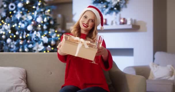 Portret van een gelukkige blanke vrouw met een glas champagne houdt de doos in de hand, schudt en glimlacht terwijl ze thuis in de buurt van een versierde kerstboom zit. Nieuwjaarsconcept. Feestdag — Stockvideo