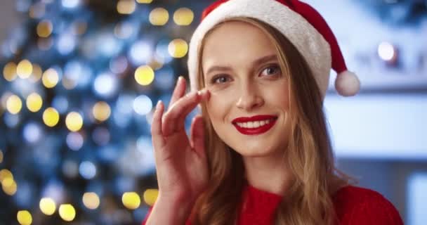 Nahaufnahme Porträt einer fröhlichen jungen schönen kaukasischen Frau mit Weihnachtsmannmütze drinnen, die in die Kamera schaut und an Silvester in positiver Stimmung lächelt. Festliche Stimmung. Frohe Feiertage — Stockvideo