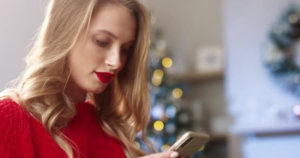 Giovane donna gioiosa caucasica in casa moderna decorata con albero di Natale che scrive e digita sullo smartphone e invia i migliori auguri con le vacanze la vigilia di Natale. Concetto spirito natalizio. Ritratto da vicino — Video Stock