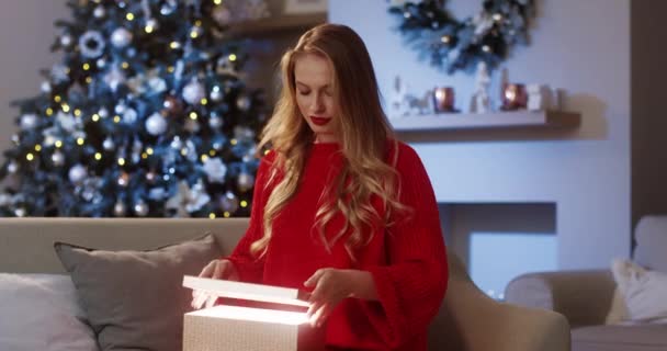 Blanke vrolijke mooie jonge vrouw in rode trui opent kerstgeschenkdoos met opgewonden verrast gezicht zich gelukkig voelen op oudejaarsavond zitten bij versierd huis in de buurt van kerstboom. Vakantieconcept — Stockvideo