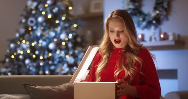 Portret radosnej białej pięknej młodej kobiety otwierającej pudełko świąteczne z podnieconą twarzą, szczęśliwej i wdzięcznej w Wigilię, siedzącej w przytulnym domu, w pobliżu świecącej choinki noworocznej. Magiczne chwile — Wideo stockowe